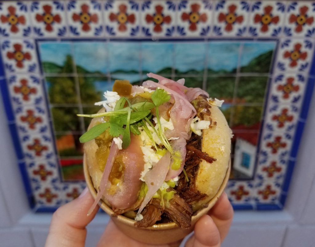 Braised Beef served on a Crispy Sourdough Roll with Queso Oaxaca, Avocado, Pickled Onions and Jalapeños in a Spicy Habanero Sauce in front of a mural in the Mexico Pavilion