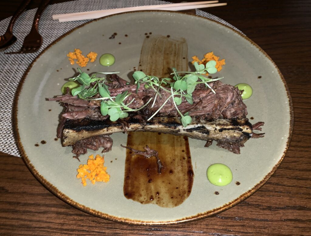 Picture of cooked, pulled meat with micro greens on top resting on a charred bone that had been cut open on a round plate with bright colored garnishings at four points around the bone and a scrape of sauce down the middle of the plate.