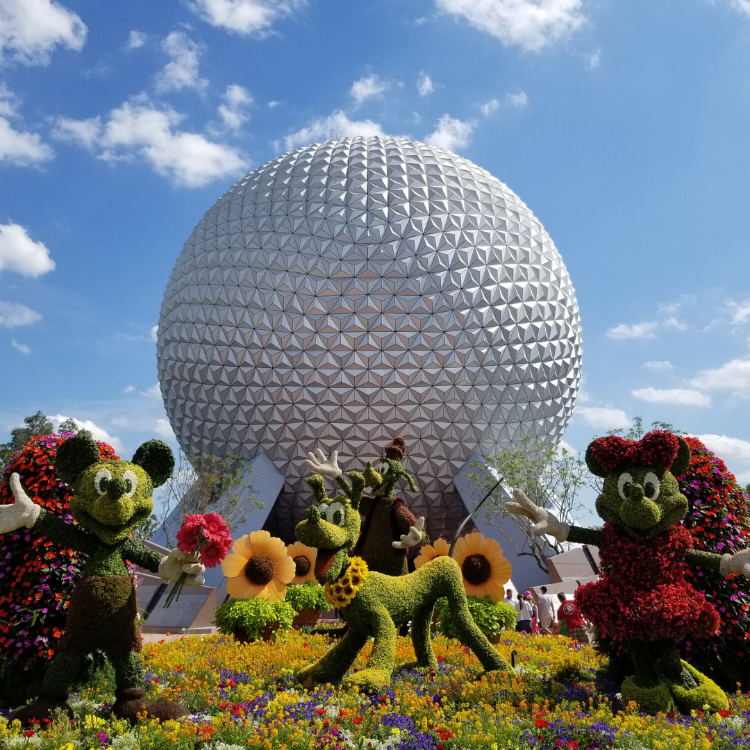 Picture of Spaceship Earth in Epcot with topiaries of Mickey Mouse, Minnie Mouse, Pluto and Goofy in the flowers in front of it on a sunny day with a few light clouds in the sky