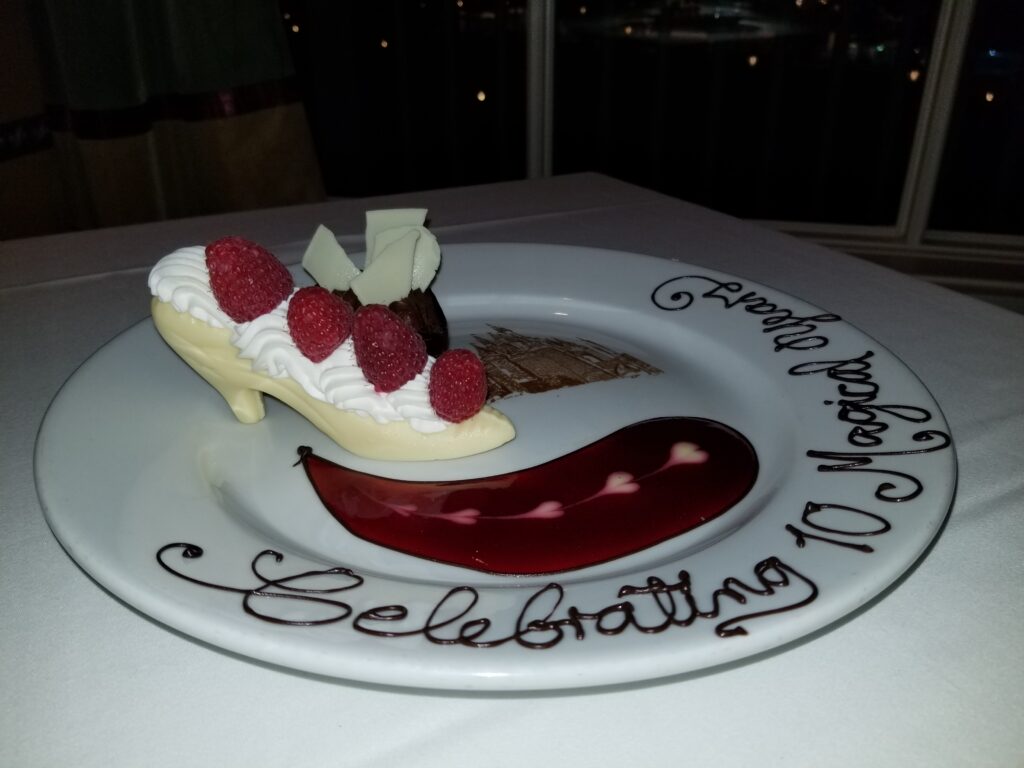 A white chocolate slipper filled with whipped cream, mouse and raspberries on a plate with a Cinderella Castle outline in chocolate and the writing, "Celebrating 10 Magical Years" 