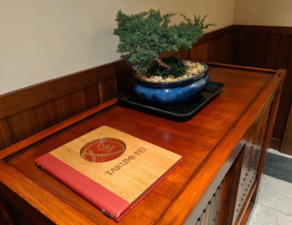 Picture of a Takumi-Tei menu and a bonsai tree on a wooden buffet.