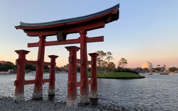 A picture of the red torii gate that is a replica of the torii gate at the Itsukushima shrine in Japan. The replica is in the World Showcase Lagoon inside Epcot in Walt Disney World at dusk with Spaceship Earth in the background.