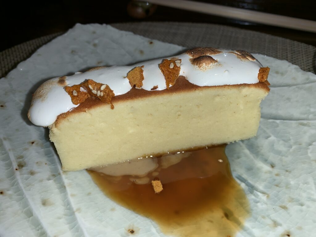 Picture of a long thing, rectangular cut of nearly pore-less cake, with a toasted meringue and small pieces of sesame brittle on top sitting over a small pool of honey caramel.
