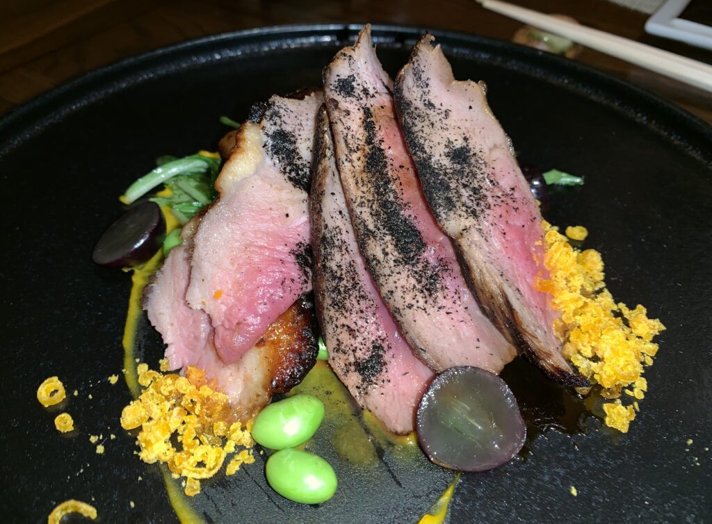 Picture of five strips of duck cooked medium rare on a plate with brightly colored garnishing and dusted with ash.