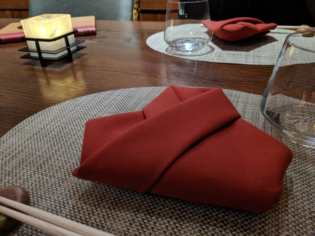 An origami napkin folded into a kimono set on a table with chopsticks in front and a dim, flickering candle nearby