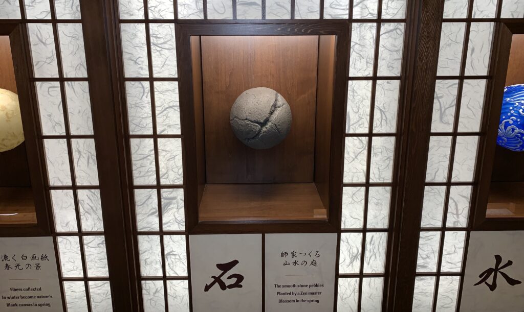 A picture of a white, granite, and blue, volleyball sized orbs that represent the elements Washi paper, stone, and water in glass cases embedded in a wall with Japanese script below.
