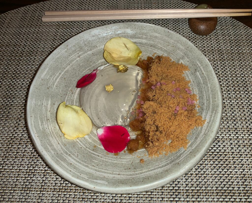 Picture of a clear and colorless dome, the size of half a softball on a plate with browned breadcrumbs to the right, two white and two red rose petals around it, and two small flakes of gold leaf on top of the water cake.