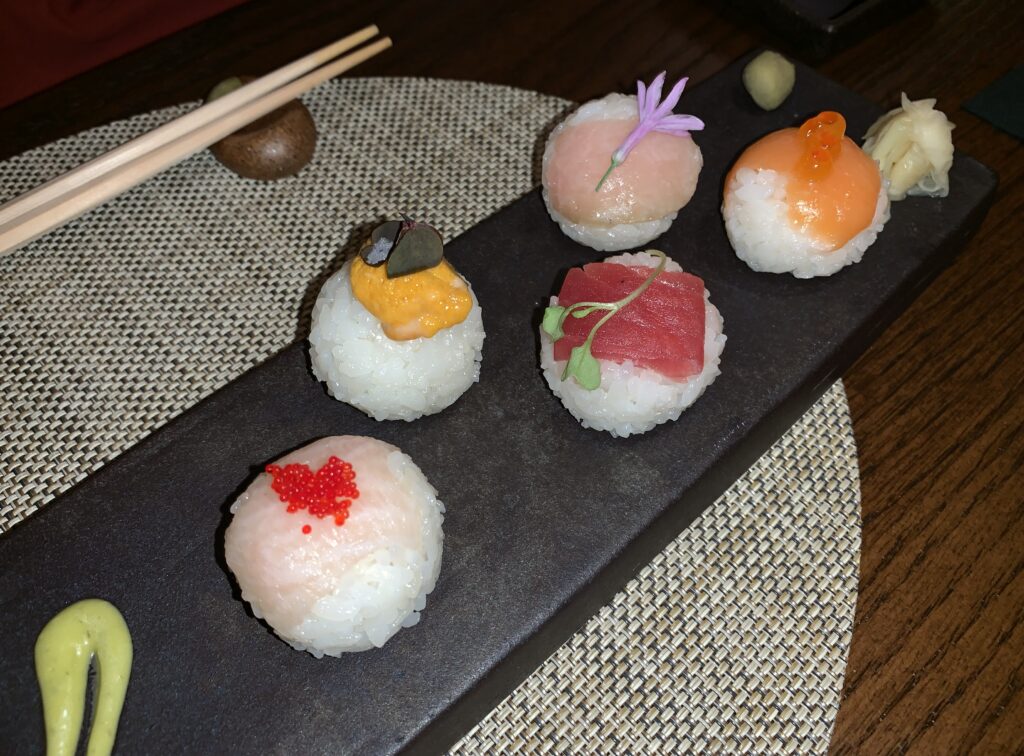 Picture of five pieces of sushi served upon a dark stone slab in a zig-zag pattern with sauces on each end.  Each thin slice of meat sits upon a golf ball sized ball of sticky rice.  From left to right, beige-colored yellow fin, intensely orange uni, red tuna, beige-colored toro, and orange salmon.