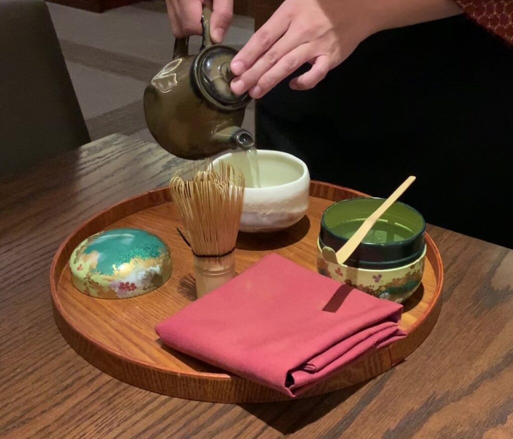 Picture of a Japanese tea set served on a wooden, circular serving tray.  Clockwise starting at 12, a white ceramic cup, a small, decorated bowl with green matcha powder in it and a serving stick resting on top, a red square folded napkin, an upright, wooden whisk, the decorated lid to the matcha bowl.  A woman’s hands are holding a small, dark olive-green tea pot pouring warm water into the white cup.