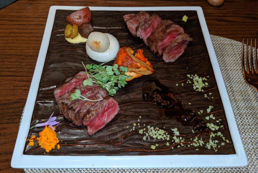 Picture of two medium-rare, sliced steaks on a large square stoneware plate garnished from upper left to lower right, three fingerling potatoes in a pale-yellow sauce, cipollini onions, orange mushrooms, a dark red Arima Sansho pepper reduction, and then a sprinkling of light green matcha maldon flakes.  From lower left to upper right, there is the bright orange yuzu kosho topped with a lavender colored garlic flower, a steak with small greens, the second steak, and then a small dollop of wasabi.