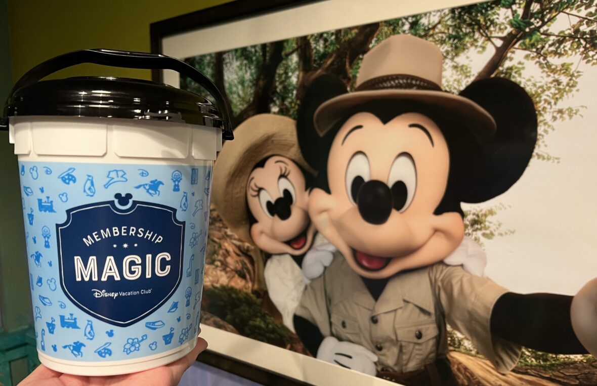 The DVC "Membership Magic" Popcorn Bucket held in front of a picture of Adventurer Mickey and Minnie Mouse at Disney's Animal Kingdom Resort.