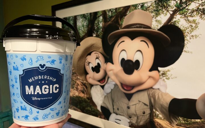 The DVC "Membership Magic" Popcorn Bucket held in front of a picture of Adventurer Mickey and Minnie Mouse at Disney's Animal Kingdom Resort.